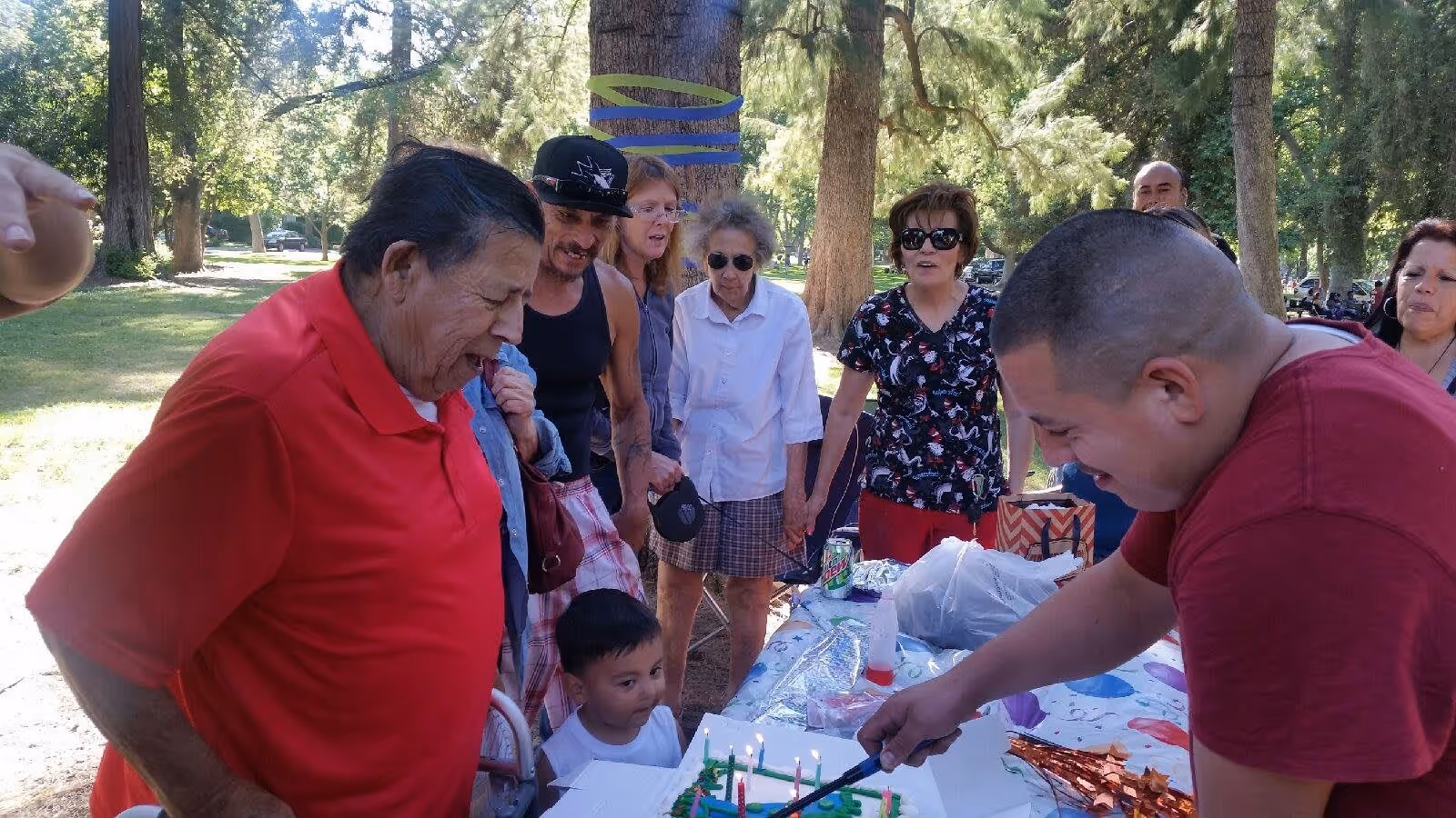 A group of people gathered outdoors around a table with a birthday cake. One man in a red shirt is blowing out candles on the cake while another man in a maroon shirt is lighting the candles. Several adults and a child are watching and standing nearby among trees in a park-like setting.