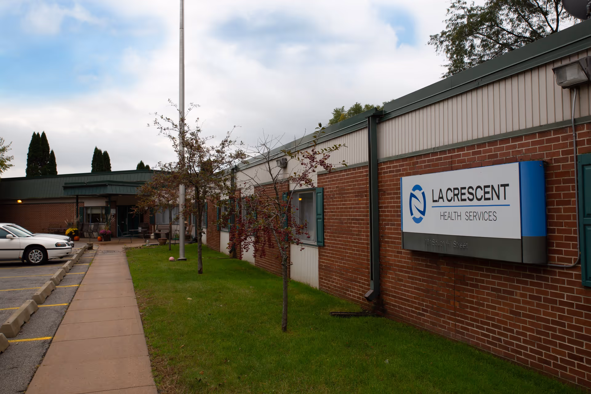 Front exterior of La Crescent Health Services brick building with a sign by a walkway, small trees, and parked cars.