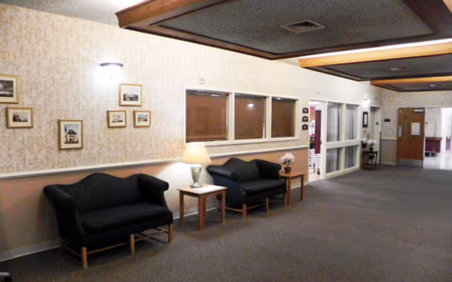 Interior hallway of a senior living facility with two black cushioned sofas, two wooden side tables with lamps and flowers, framed pictures on the wall, and a carpeted floor. The ceiling has wooden beams and recessed lighting.