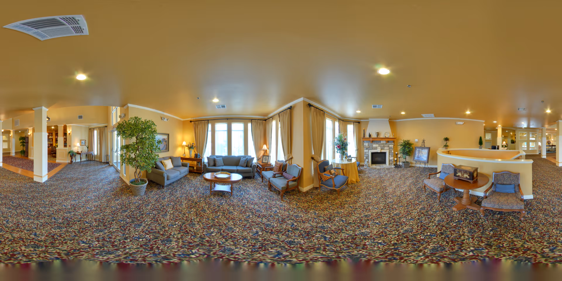 A spacious and well-lit living room area in a senior living facility with patterned carpet, multiple seating arrangements including sofas and armchairs, a coffee table, potted plants, large windows with curtains, and a stone fireplace with decorative items on the mantel.