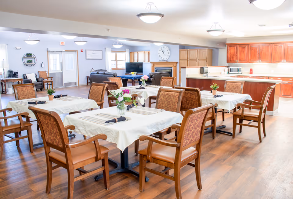 A bright and spacious dining area in a senior living facility featuring several tables covered with white tablecloths, each set with napkins and small flower arrangements. The room has wooden chairs with cushioned seats, hardwood floors, and a kitchen area with wooden cabinets and appliances in the background. There is also a seating area with sofas and a TV near a fireplace.