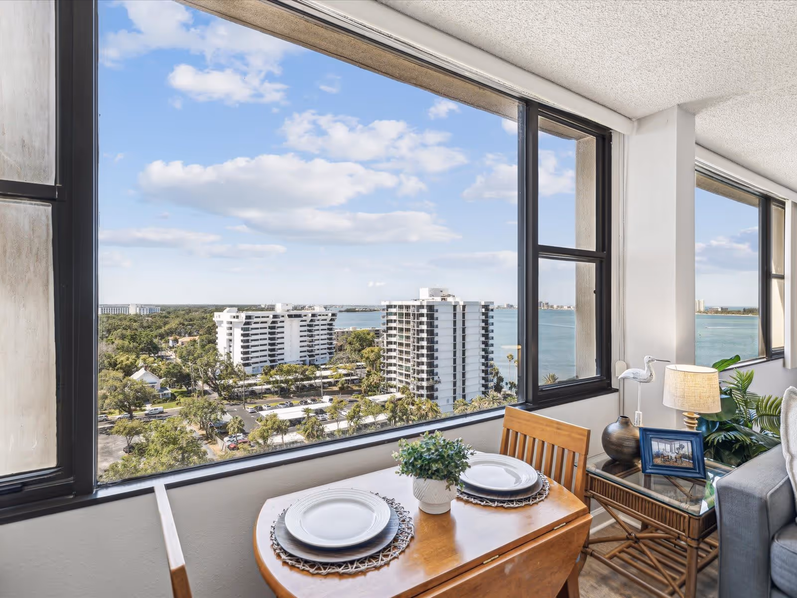 A bright interior space with a small wooden dining table set for two with white plates and a small potted plant in the center. Next to the table is a glass-top side table with a lamp, a decorative bird figurine, and a framed photo. Large windows provide a panoramic view of nearby buildings, greenery, and a body of water under a partly cloudy sky.