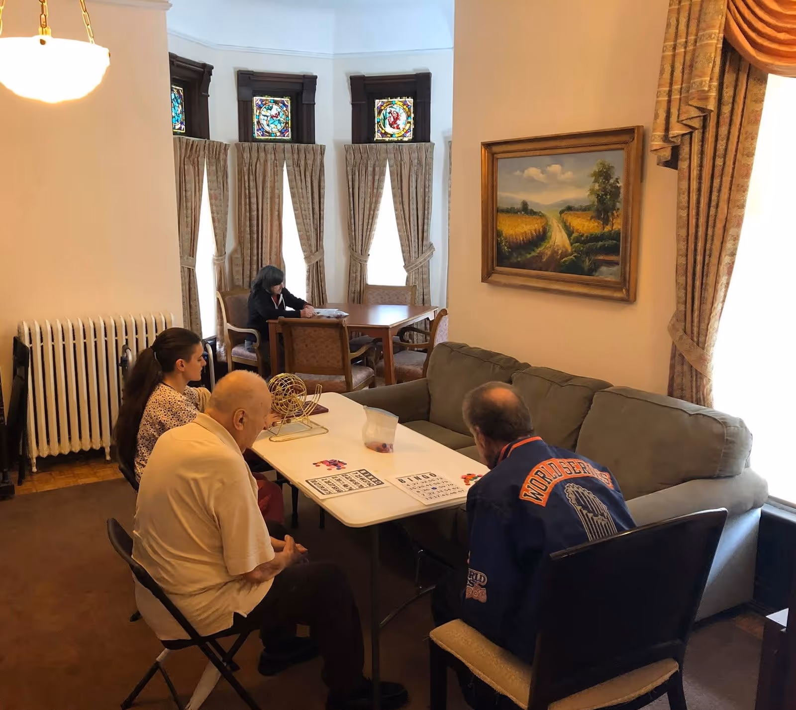Three elderly people sitting around a table playing bingo in a cozy room with a couch, a painting on the wall, and large windows with curtains and stained glass panels. Another person is sitting at a separate table in the background.