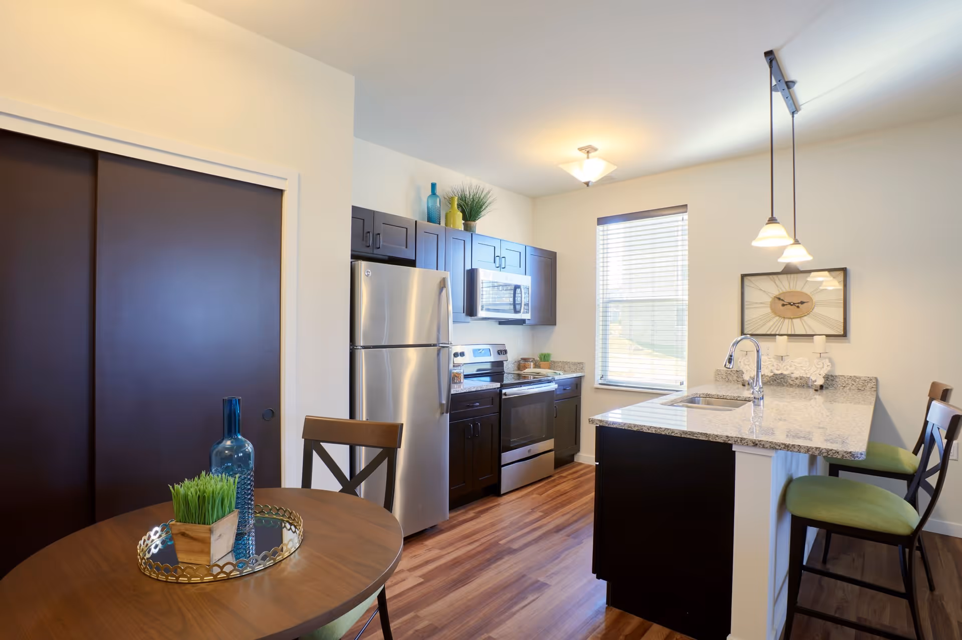Modern kitchen with dark wood cabinets, stainless steel refrigerator, stove, and microwave. There is a granite countertop island with a sink and three pendant lights hanging above it. A round wooden dining table with two chairs is in the foreground, decorated with a blue bottle and a small plant. A window with blinds allows natural light into the room.