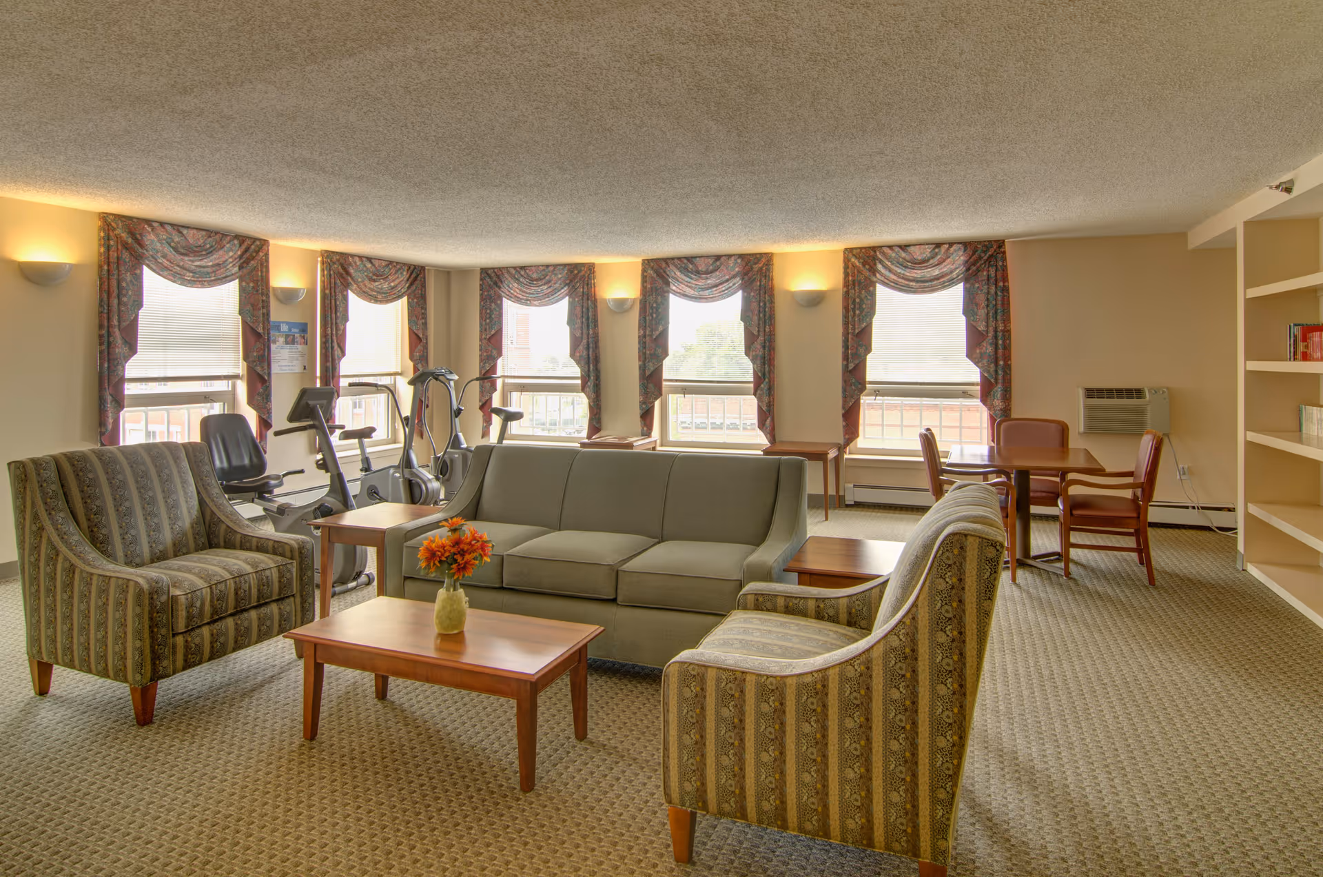 A well-lit common area with patterned armchairs, a green sofa, wooden coffee tables, and exercise equipment near windows with floral valances. There is also a small table with chairs and built-in shelves on the right side of the room.