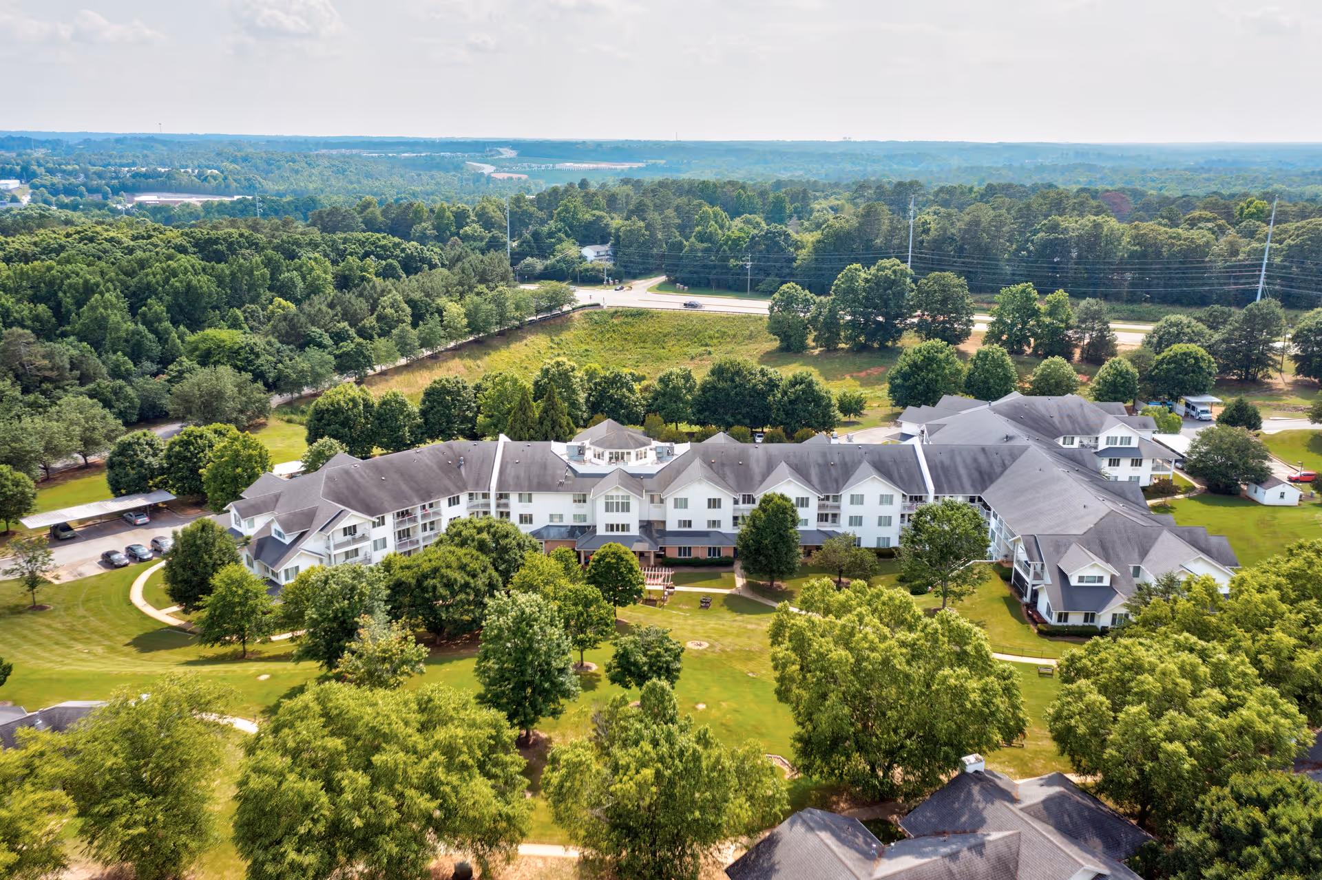 Aerial view of TerraBella Epps Bridge senior living facility surrounded by green trees and lawns, with a large white multi-wing building and a parking area visible nearby.