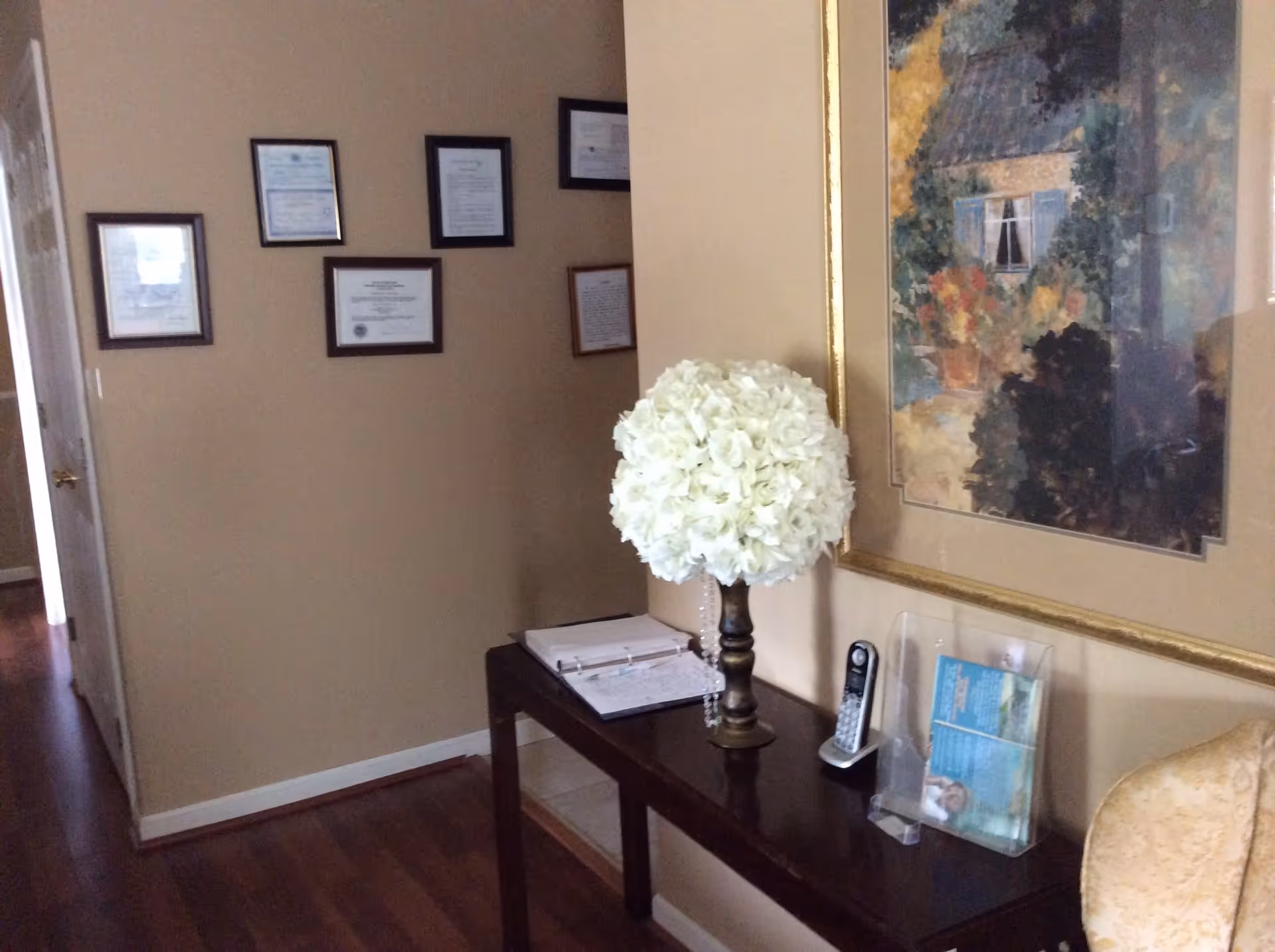 Interior hallway area with a dark wooden console table holding a white floral arrangement, a cordless phone, and a clear brochure holder. The wall behind the table features a large framed painting of a house surrounded by greenery and flowers. On the adjacent wall, several framed certificates or documents are displayed. The floor is wooden, and a partially visible door is on the left side.