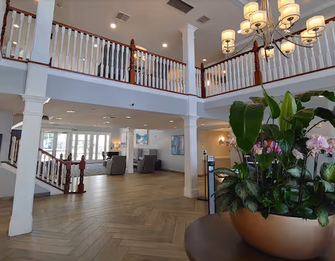 Spacious interior of a senior living facility with a two-story ceiling, white columns, wooden railings on the upper level, a large chandelier with multiple lights, a large potted plant with green leaves and pink flowers, and seating areas with chairs and lamps in the background near large windows.