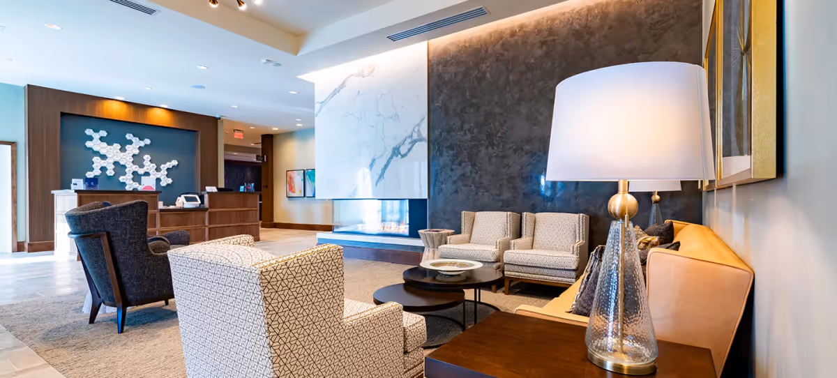 A modern and elegant senior living facility lobby with comfortable seating including patterned armchairs and a tan sofa. There is a large marble fireplace with a dark textured wall behind it. A wooden reception desk is visible in the background with decorative wall art above it. A glass table lamp with a white shade is on a side table next to the sofa.