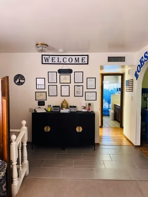 Interior view of a welcoming area in a senior living facility with a black cabinet against a white wall. Above the cabinet is a large 'WELCOME' sign and several framed certificates and documents arranged neatly. To the left is a white staircase railing, and to the right is an open doorway leading to another room with wooden flooring and a glimpse of a bathroom. The floor in the foreground is tiled.