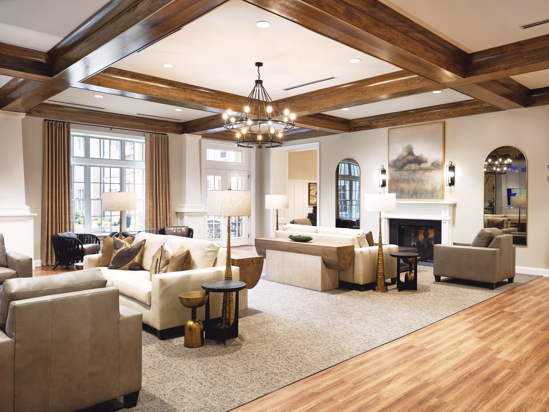 A spacious senior living facility common area with beige and gray sofas and armchairs arranged around a fireplace. The room features wooden ceiling beams, a large chandelier, floor lamps, and large windows with brown curtains letting in natural light. There are decorative mirrors and artwork on the walls, and the floor is a combination of wood and carpet.