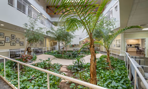 Indoor atrium area with lush green plants and small trees surrounded by a white railing. The space is located inside a two-story building with a high ceiling and large windows allowing natural light. Benches and framed pictures line the walls on the left side, and a seating area is visible on the right. The overall atmosphere is bright and inviting.