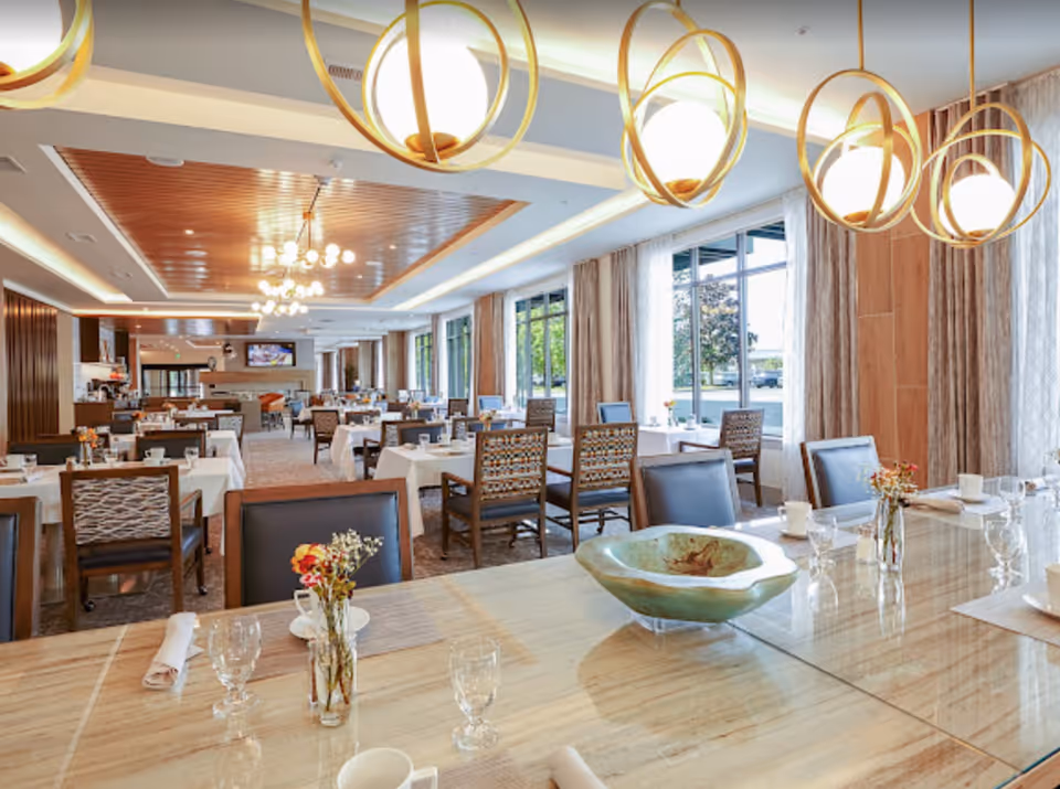 Bright and spacious dining room with multiple tables covered in white tablecloths, set with glasses, cups, and napkins. Modern pendant lights hang from the ceiling, and large windows allow natural light to fill the room. Chairs with patterned backs and leather seats are arranged around the tables, and a decorative bowl and small flower vases are placed on the foreground table.