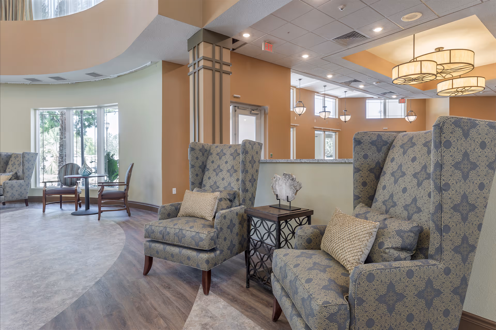 A bright and spacious senior living facility lounge area with patterned wingback chairs, a small side table with decorative art, large windows letting in natural light, and modern hanging light fixtures.