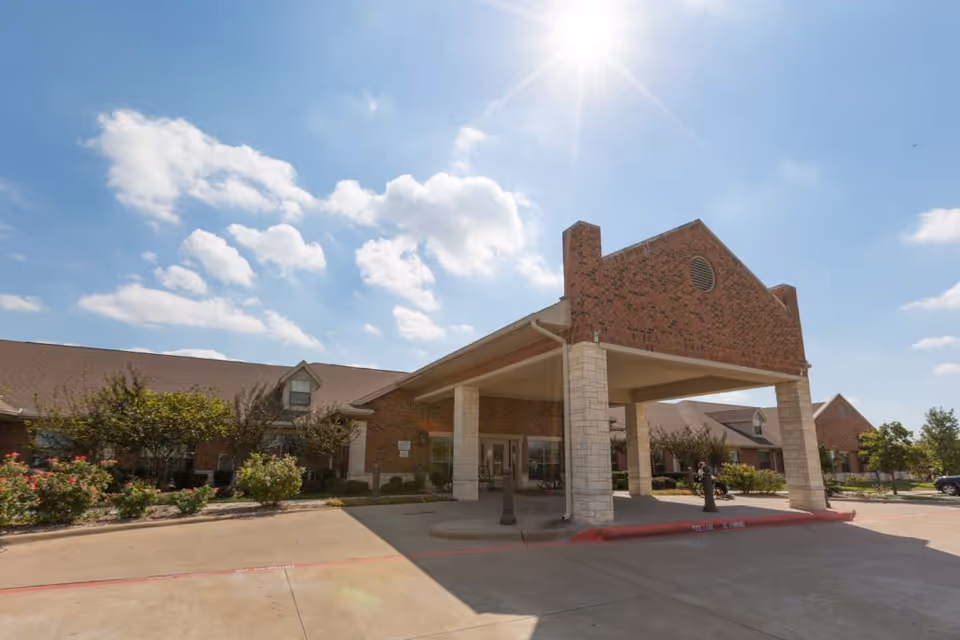 Exterior view of Legend Oaks Healthcare and Rehabilitation - Ennis building with a covered entrance supported by stone pillars, surrounded by landscaping and a clear blue sky with some clouds.
