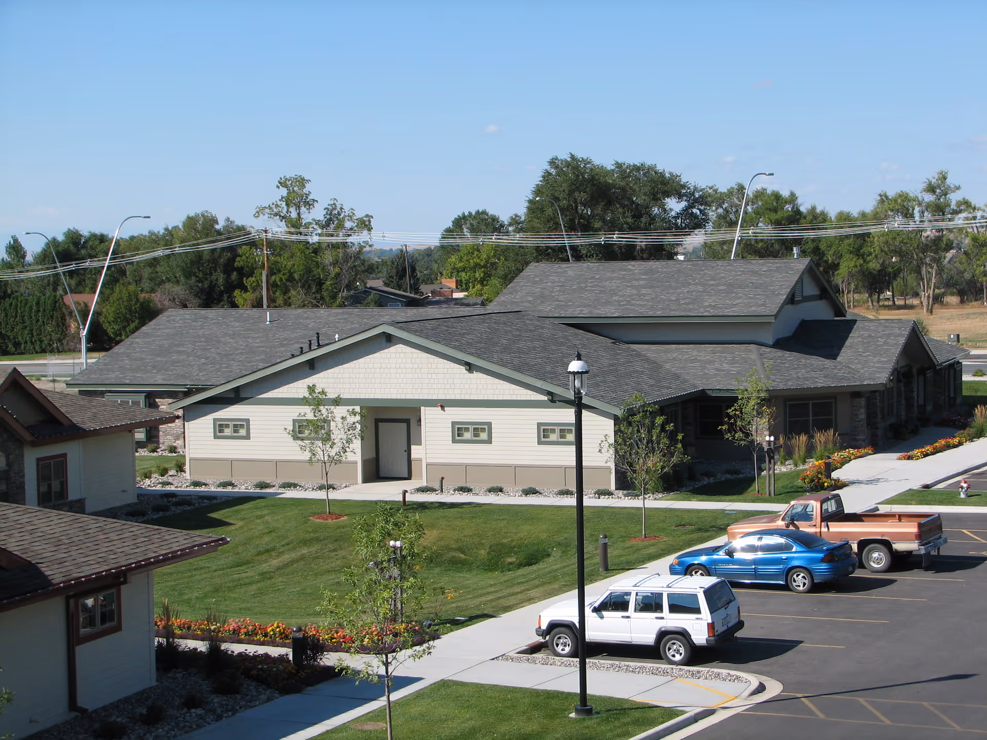 Exterior view of a single-story building with a gray roof and light-colored walls, surrounded by green lawns, small trees, and flower beds. Several cars are parked in the parking lot in front of the building under a clear blue sky.