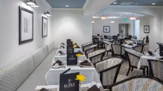 Well-lit dining room with rows of neatly set tables, upholstered chairs, menus, napkins, and small yellow flowers.