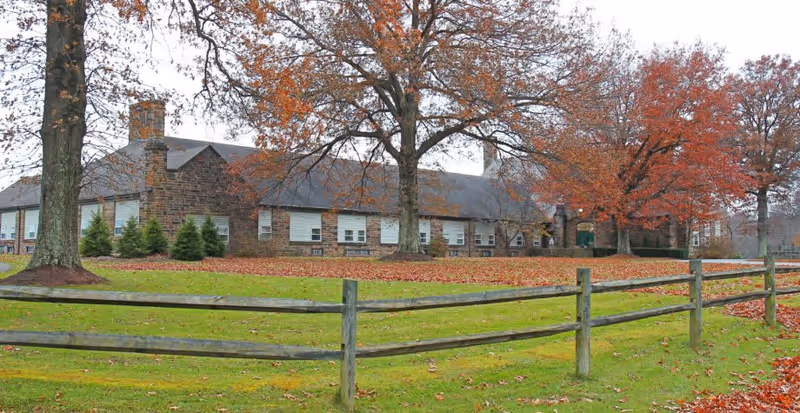 A large stone building with multiple windows surrounded by a grassy area with fallen autumn leaves and several trees with orange and red foliage. A wooden fence runs along the foreground.
