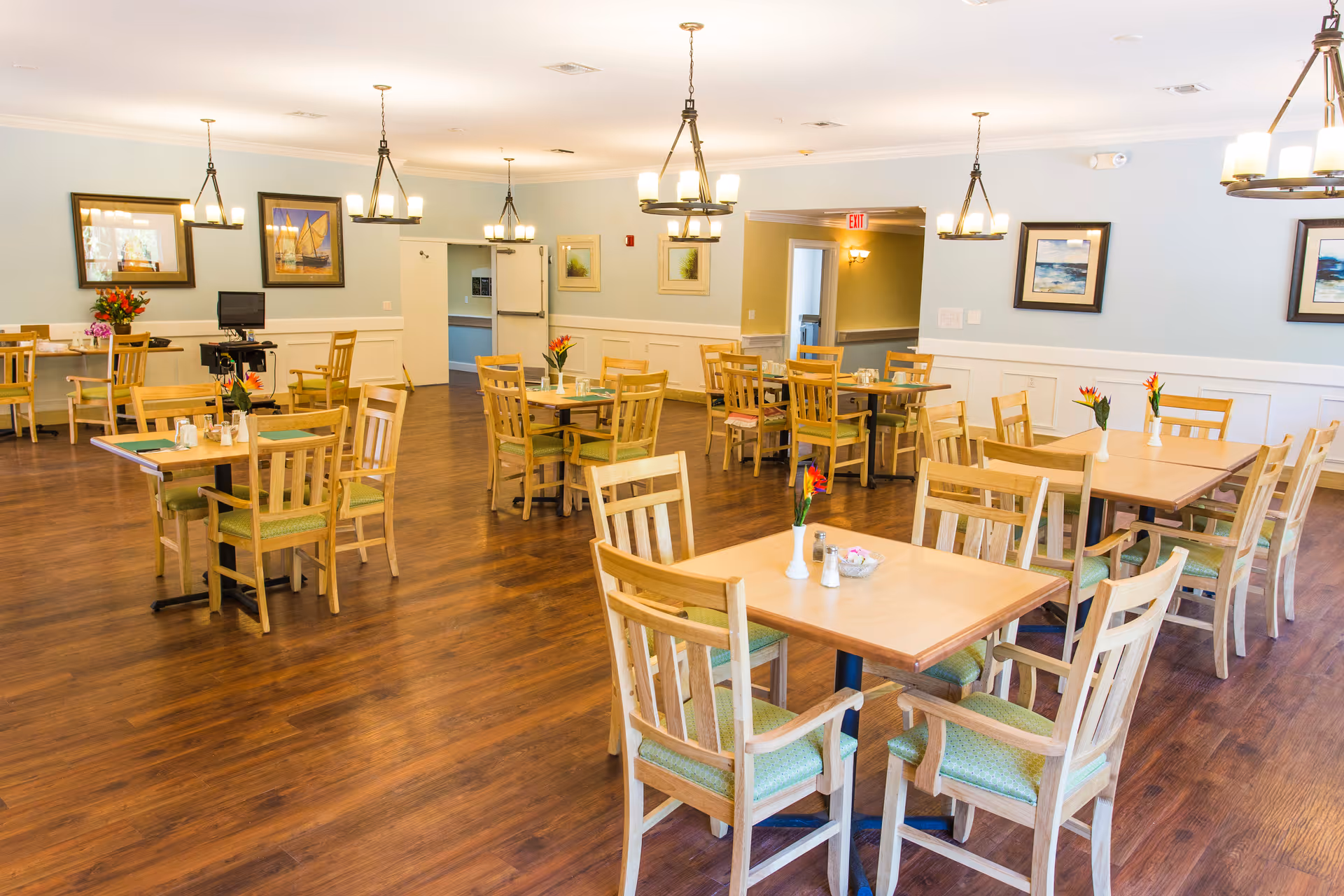 A bright and spacious dining room with several wooden tables and chairs arranged neatly. Each table has a small vase with flowers and salt and pepper shakers. The room has wooden flooring, light blue walls with white wainscoting, framed artwork on the walls, and multiple hanging light fixtures providing warm lighting.