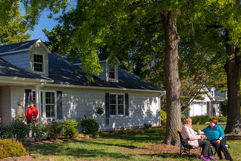 Two elderly women sitting on a bench under large trees in a grassy outdoor area, engaged in conversation. In the background, two other elderly women stand near the entrance of a white house with black shutters, surrounded by bushes and greenery.