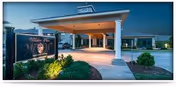 Entrance of Wilshire Place assisted living facility at dusk, showing a covered drop-off area with white columns, well-lit exterior, and landscaped greenery including bushes and mulch beds.