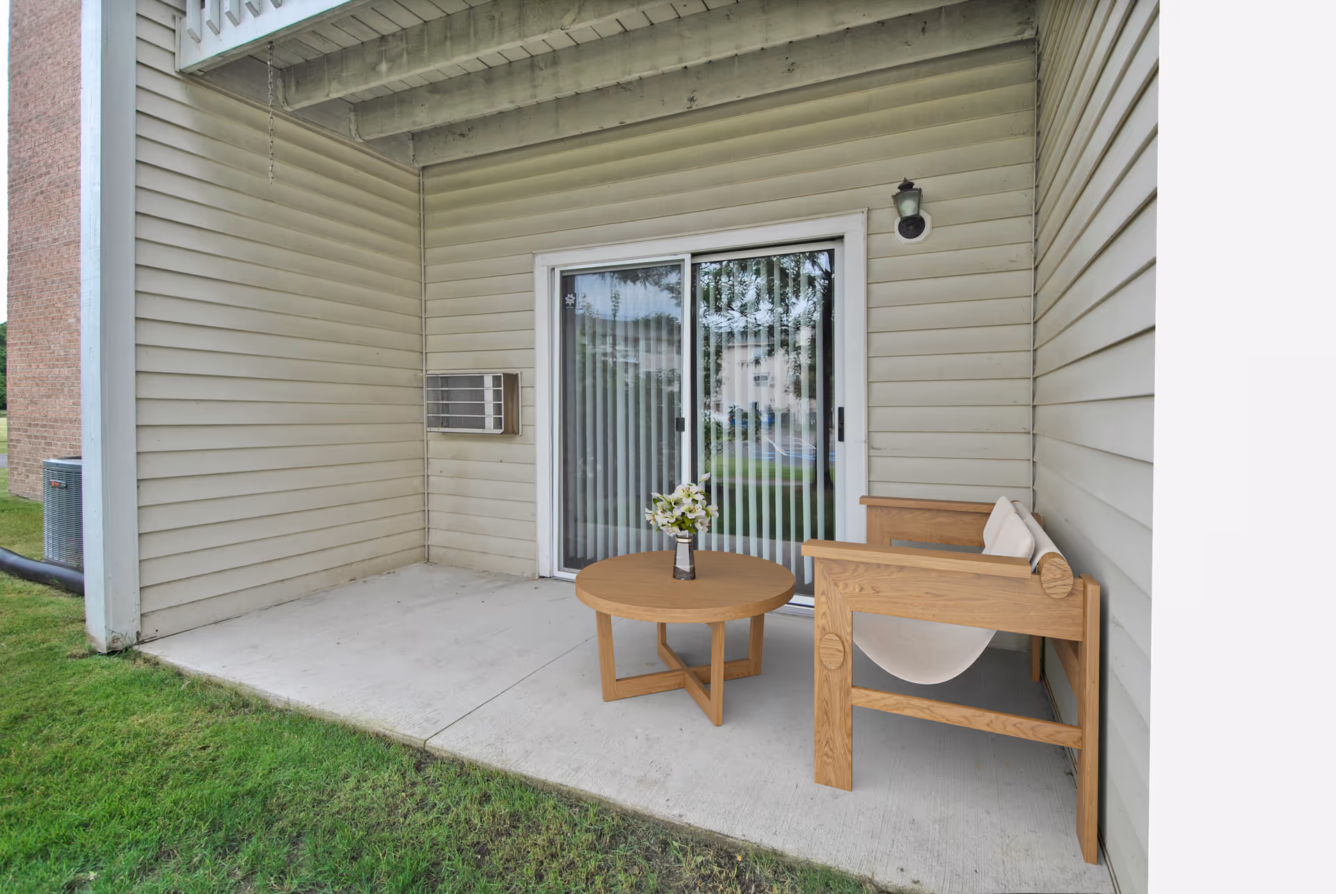 Covered outdoor patio area with beige siding walls, a sliding glass door with vertical blinds, a wooden chair with white cushions, and a round wooden table with a vase of white flowers on it. Green grass surrounds the concrete patio.
