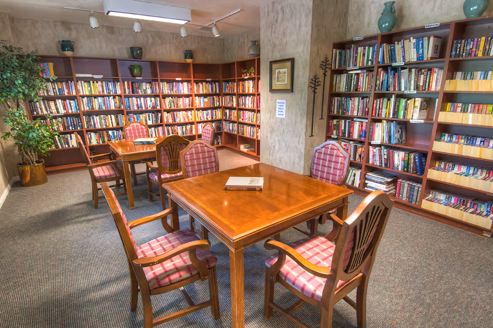 Well-lit library/reading room with bookshelves lining the walls and wooden tables with upholstered chairs.
