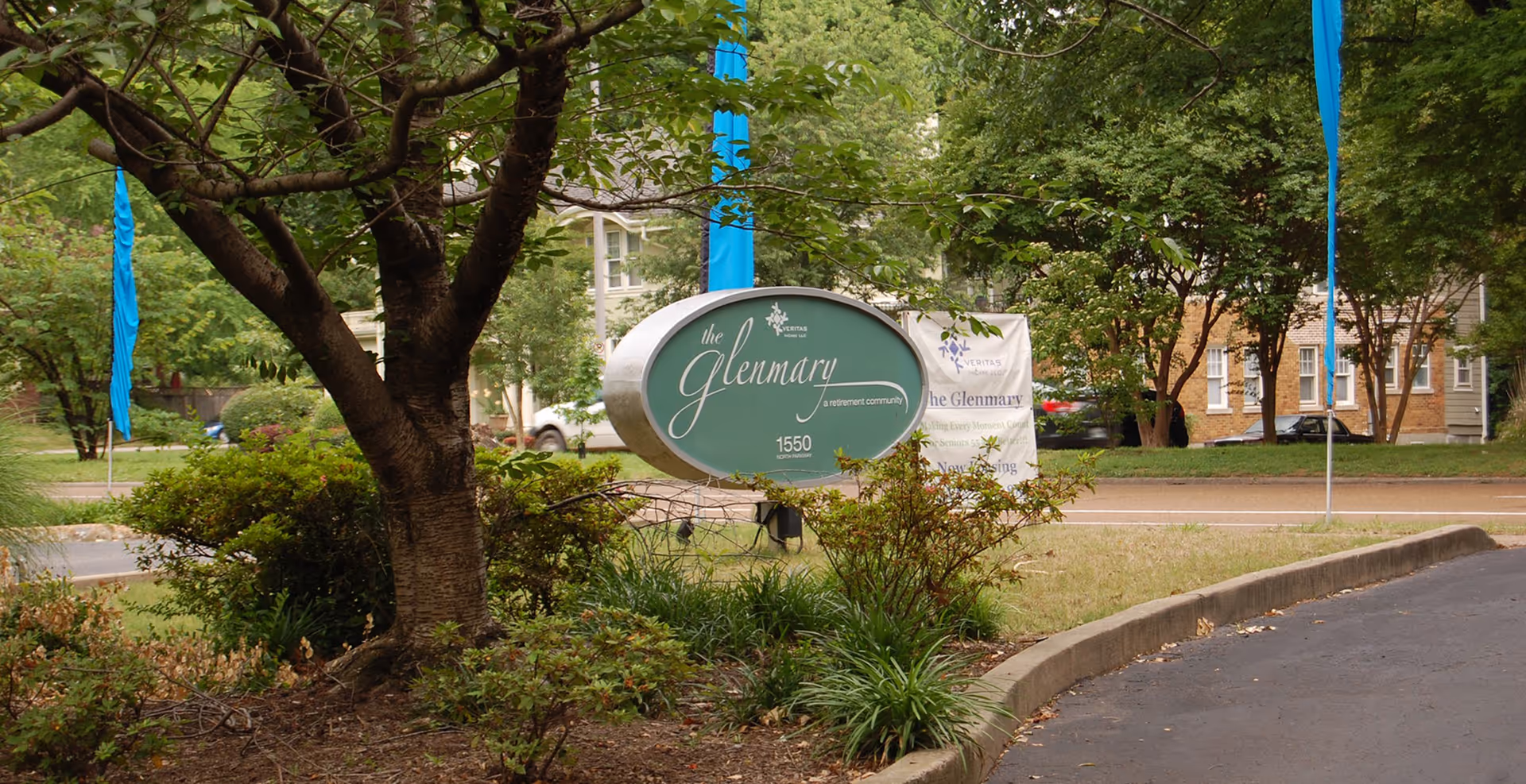 Outdoor view of the entrance area to The Glenmary retirement community, featuring a green oval sign with the community's name and address surrounded by trees, bushes, and a paved driveway.