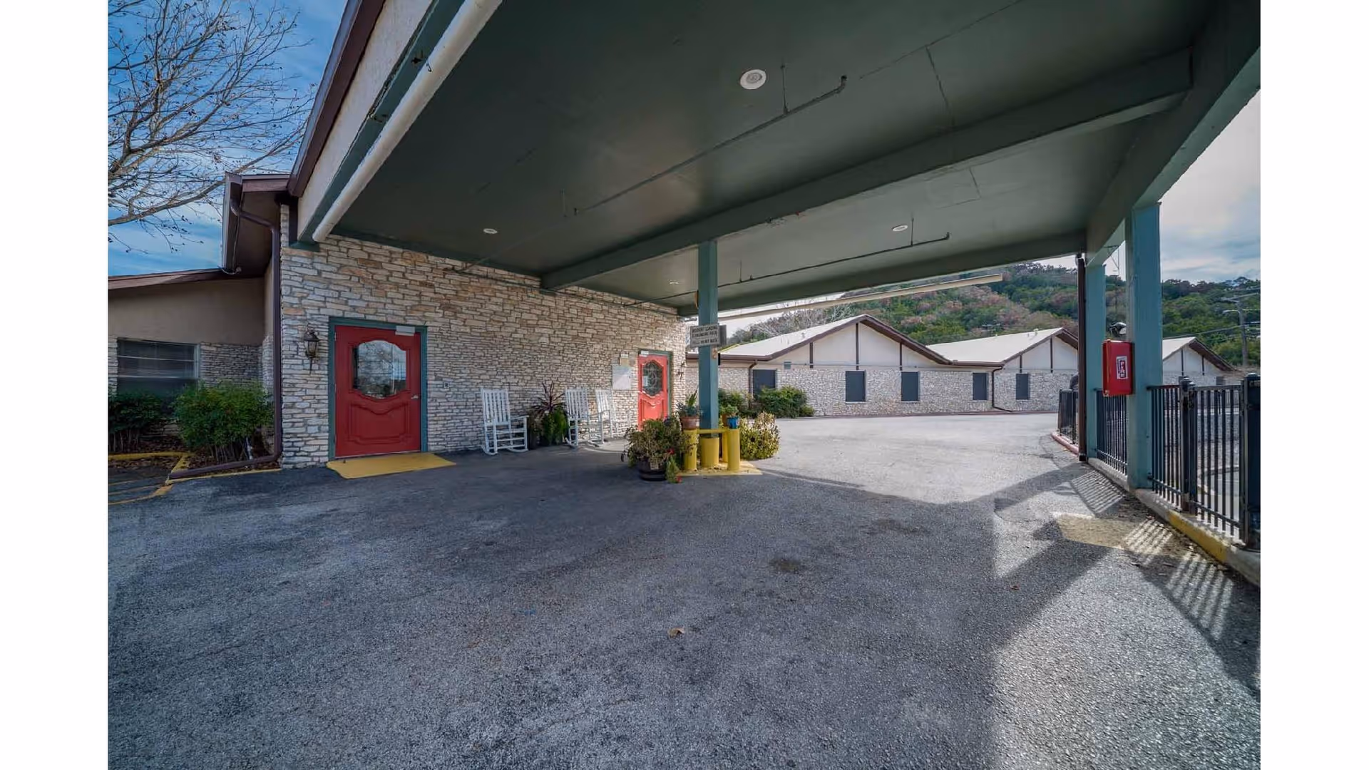 Covered porte-cochere and driveway at the front entrance of a stone-faced nursing facility with red doors and a pair of white rocking chairs.