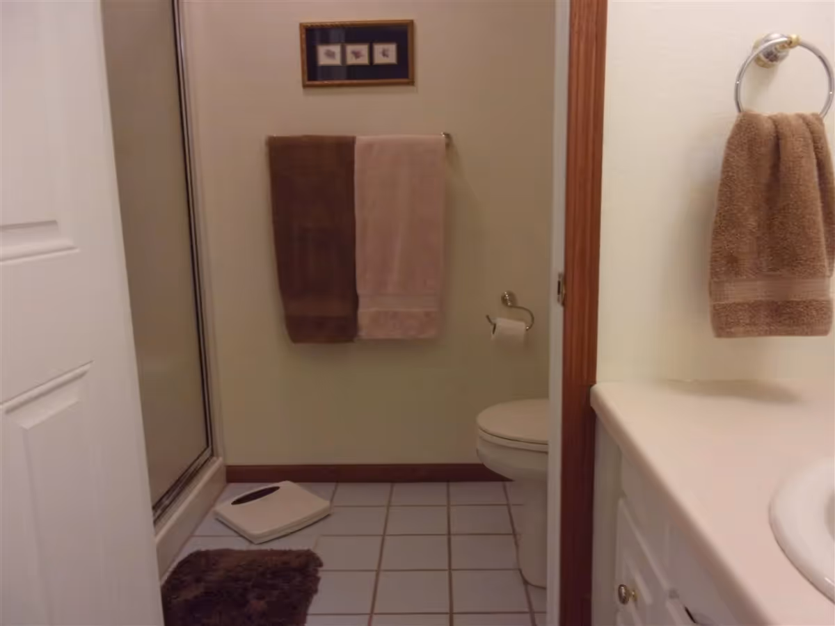 A bathroom with a shower on the left, a toilet on the right, and two towels hanging on a rack on the wall. There is a brown bath mat on the tiled floor and a white scale next to the shower. A brown hand towel hangs on a ring near the sink countertop.