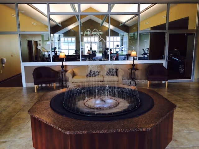 Indoor seating area with a central water fountain surrounded by a brown octagonal countertop. Behind the fountain, there are two armchairs on either side of a beige sofa with two side tables and lamps. The background features large windows looking into another room with exercise equipment and a high ceiling with exposed beams.