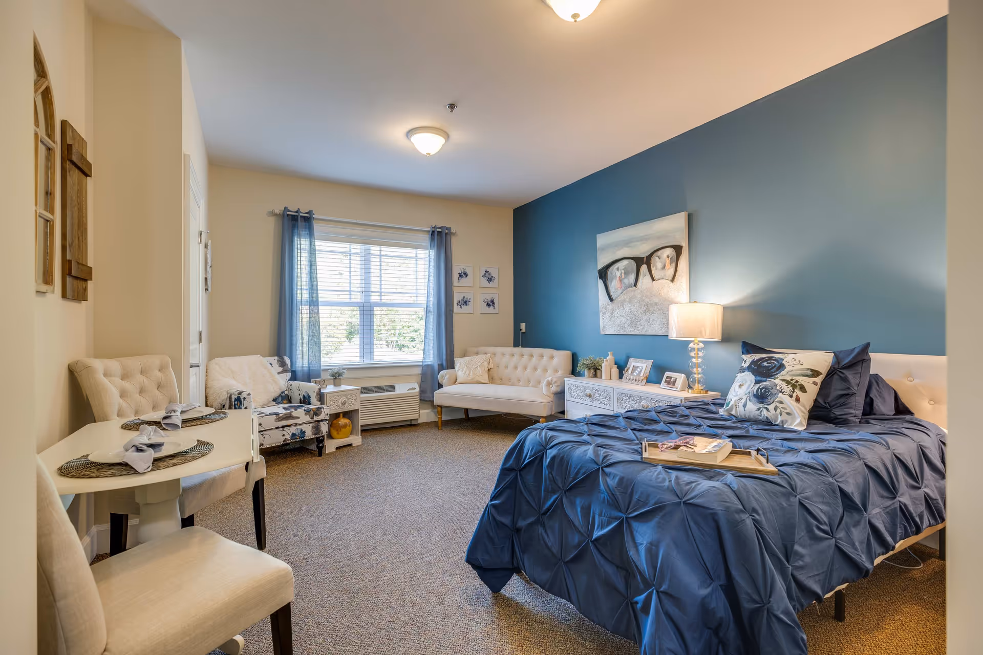 A cozy bedroom in an assisted living facility with a blue accent wall behind the bed. The bed is made with a blue comforter and floral pillow. There is a bedside table with a lamp and decorative items. The room also includes a small dining table with two chairs, a cushioned armchair with a floral pattern, a small white loveseat, and a window with blue curtains letting in natural light.