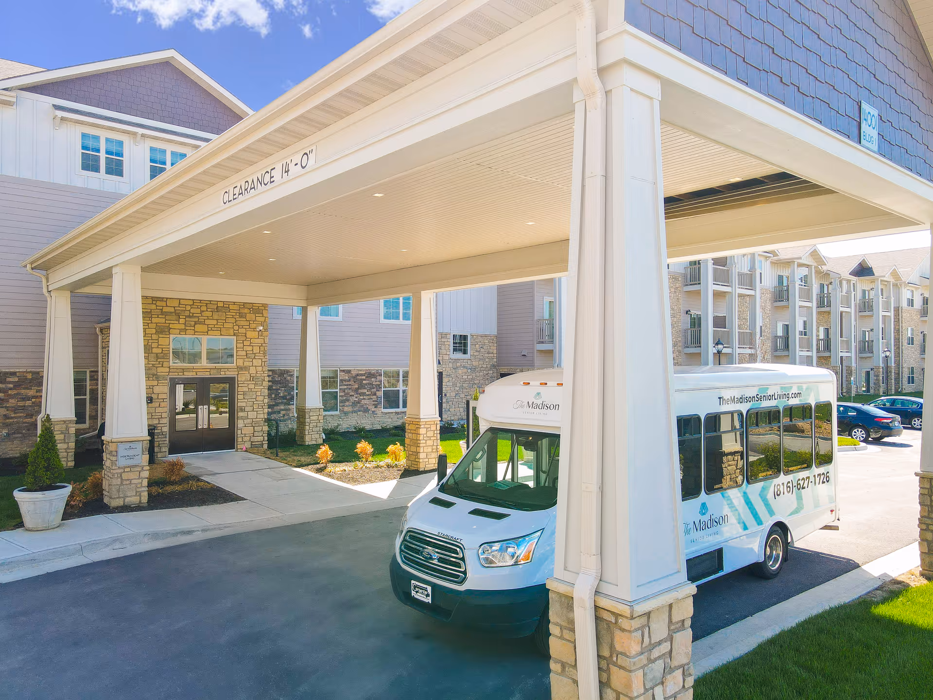 Entrance of The Madison Senior Living facility with a covered drop-off area. A white shuttle bus with The Madison Senior Living branding and contact information is parked under the canopy. The building exterior features stone and siding with multiple windows and balconies. Cars are parked in the background under a clear blue sky.