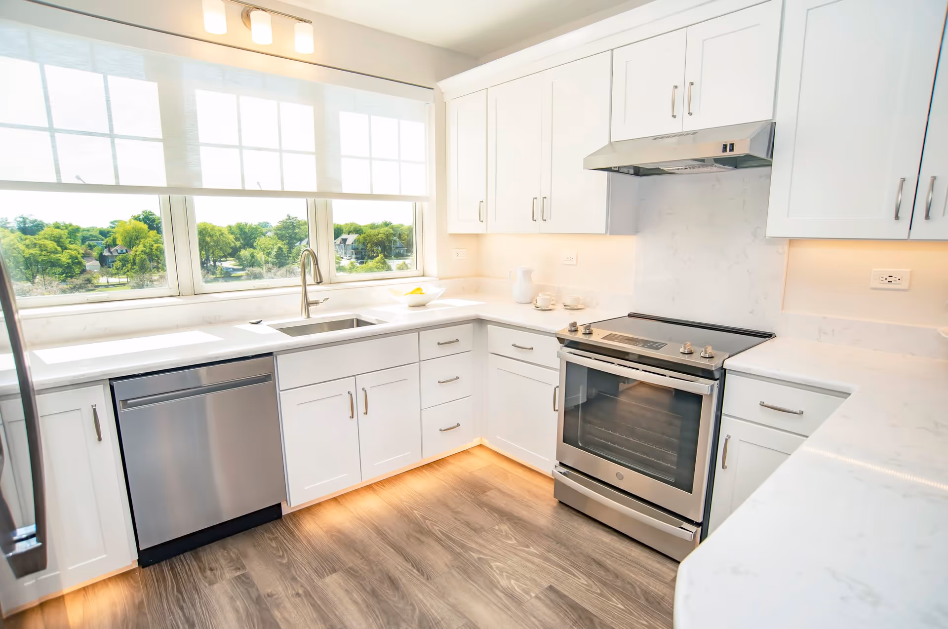Bright modern kitchen with white cabinets, stainless steel appliances including a dishwasher and oven, a large window with a view of green trees, and wood flooring.