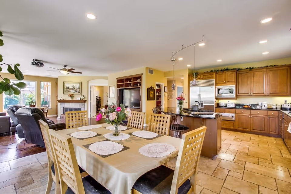 A bright and spacious open-concept dining and living area in a senior living facility. The dining area features a rectangular table with six wooden chairs and a beige tablecloth with placemats and a vase of flowers. The living area has leather recliners, a fireplace with a framed painting above it, and a large TV set in a wooden entertainment center. The kitchen area has wooden cabinets, a large island with bar stools, stainless steel appliances, and pendant lighting. The floor is tiled and the walls are painted a soft yellow.