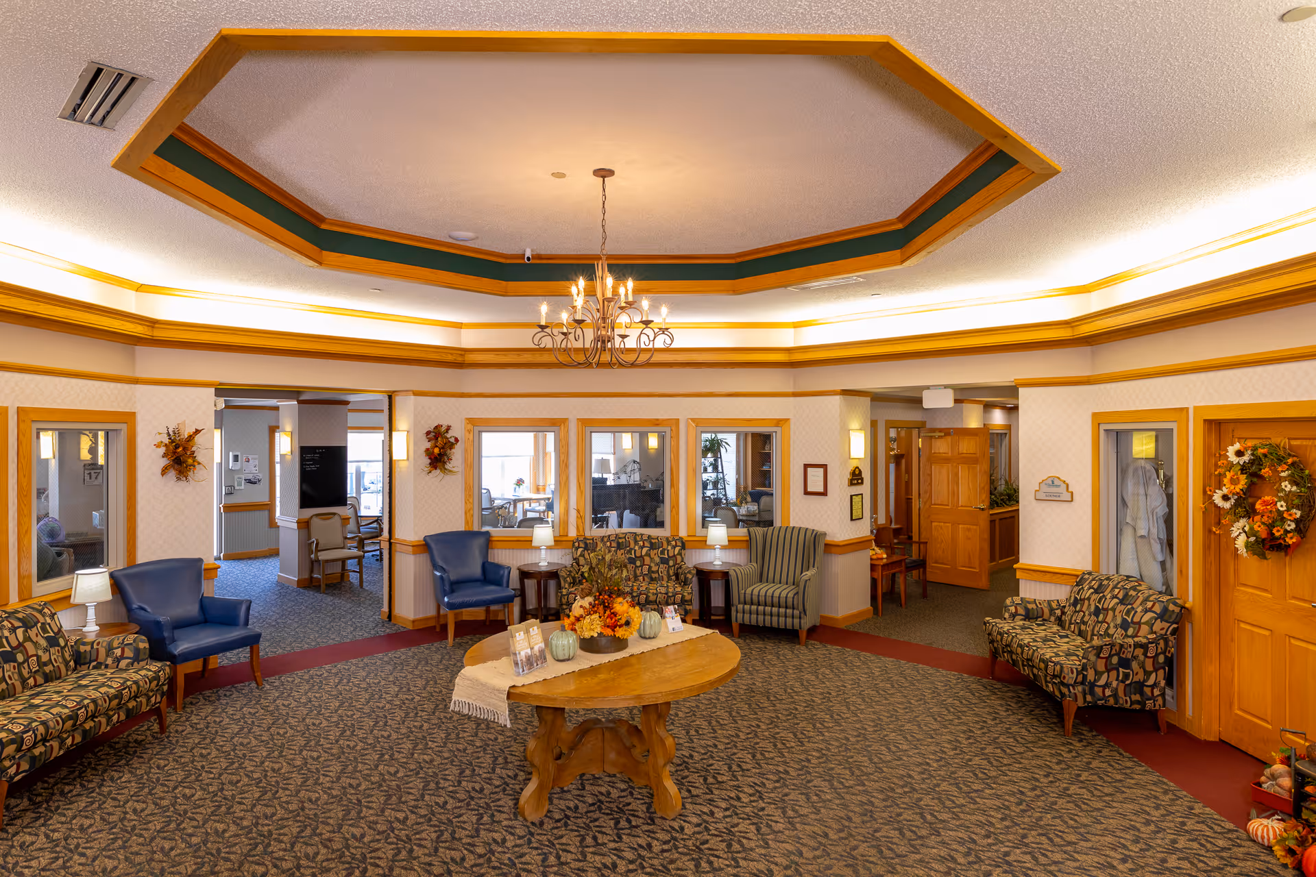A spacious and warmly decorated assisted living facility common area with patterned carpet, multiple upholstered chairs and sofas, a wooden round table with autumn-themed decorations, and a chandelier hanging from a recessed ceiling. The walls have wood trim and there are several doorways and windows leading to other rooms.