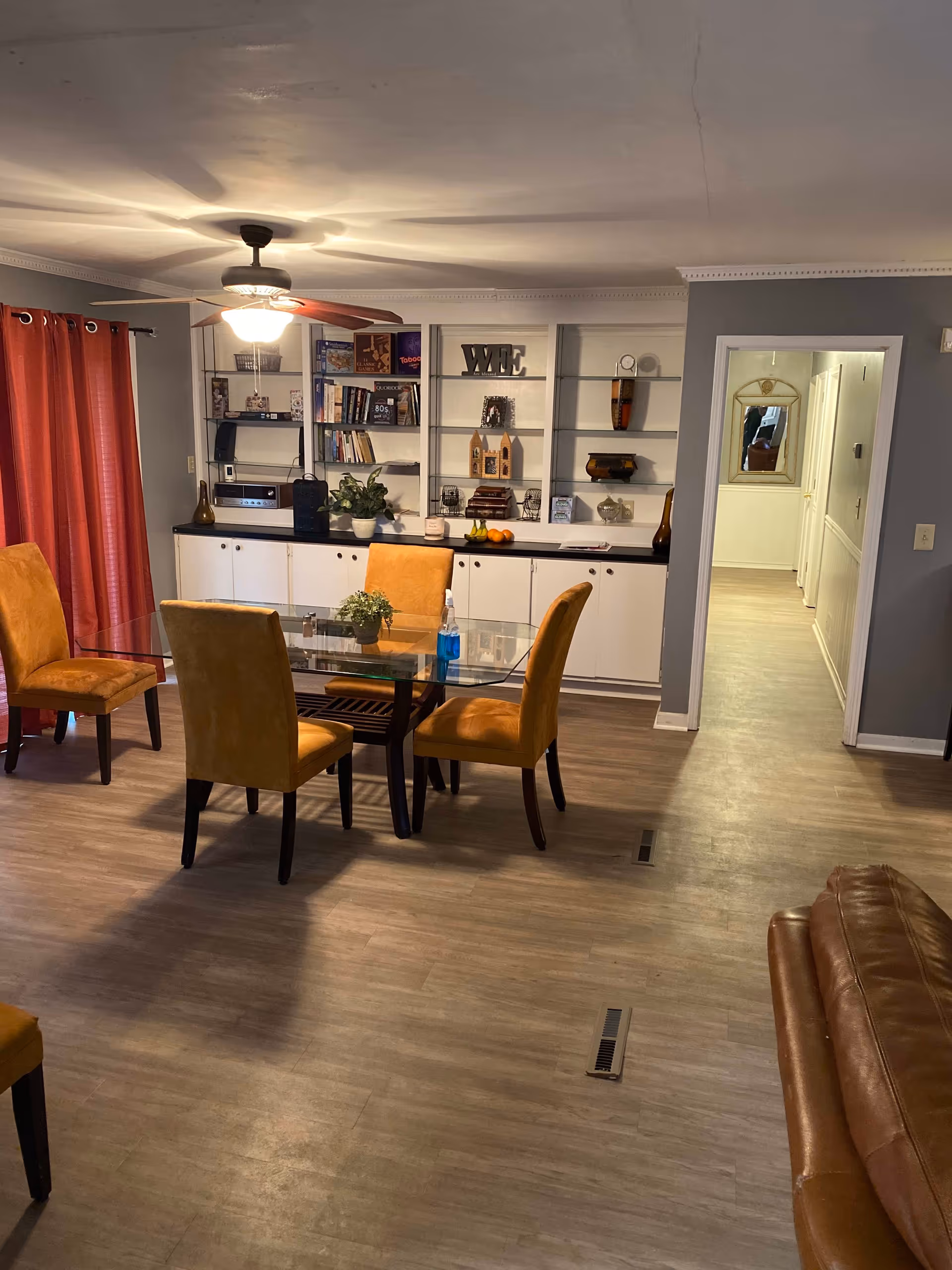 A dining area with a glass-top table surrounded by four mustard yellow upholstered chairs. Behind the table is a built-in white cabinet with shelves displaying books, decorative items, and plants. A ceiling fan with a light is on the ceiling, and red curtains cover a window on the left side. The floor is a light wood laminate, and a hallway is visible in the background.