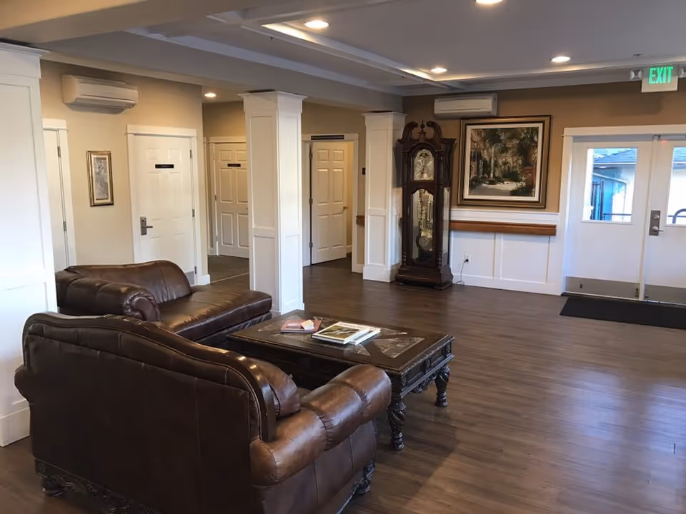 A spacious and well-lit common area with two brown leather sofas facing a wooden coffee table with books on it. The room has wooden flooring, white paneled walls, and several white doors. A large grandfather clock stands against one wall beneath a framed painting. There is an exit door with windows on the right side and an illuminated green exit sign above it.