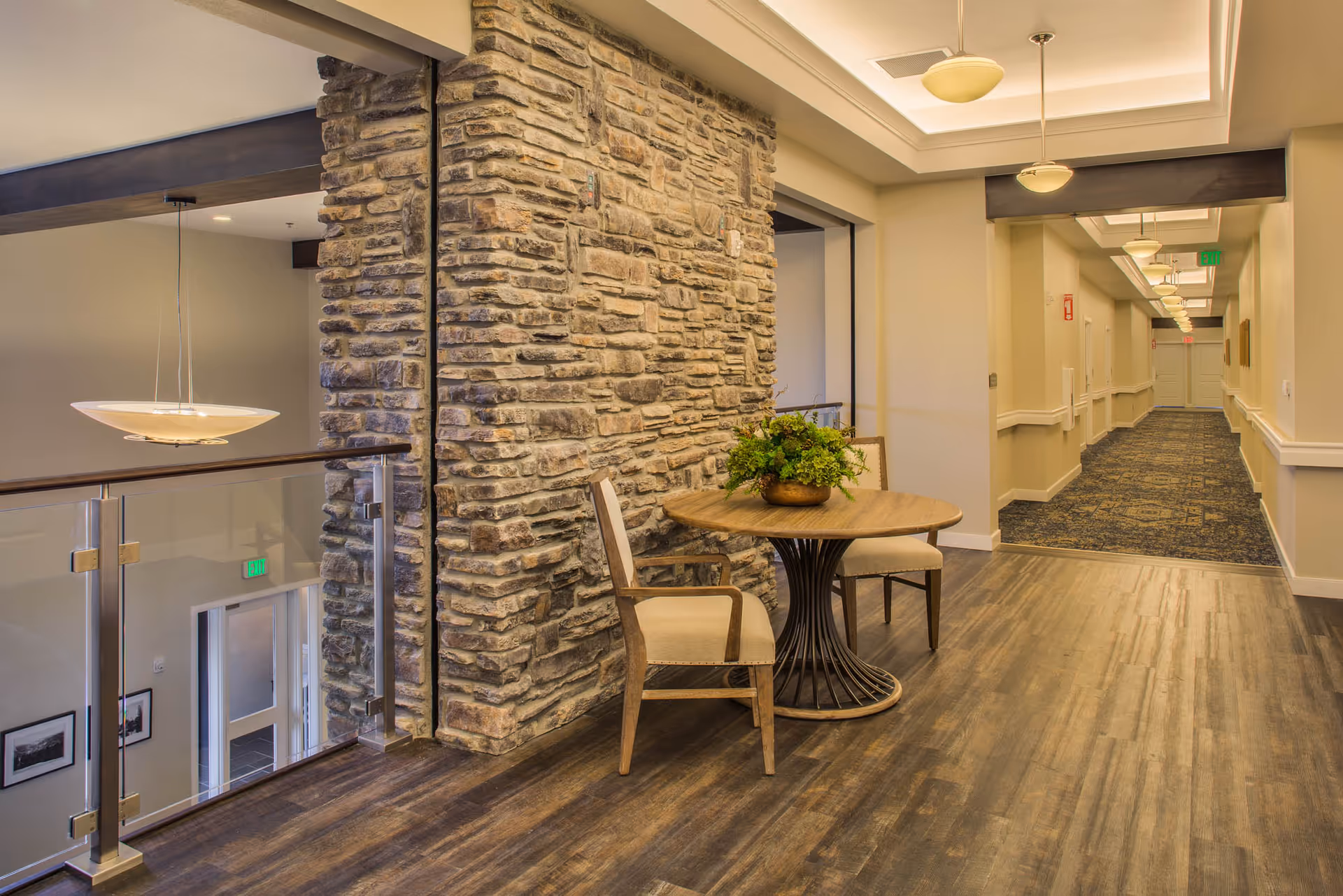 Upstairs interior sitting area with a small round table and two chairs beside a stone column overlooking a hallway and stairwell.