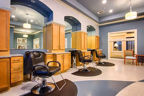 Interior view of a salon area with three black salon chairs in front of wooden cabinets and large mirrors. The walls are painted blue with white trim, and there is a window with a chair and cushion nearby. The floor has a circular pattern with dark mats under each salon chair.