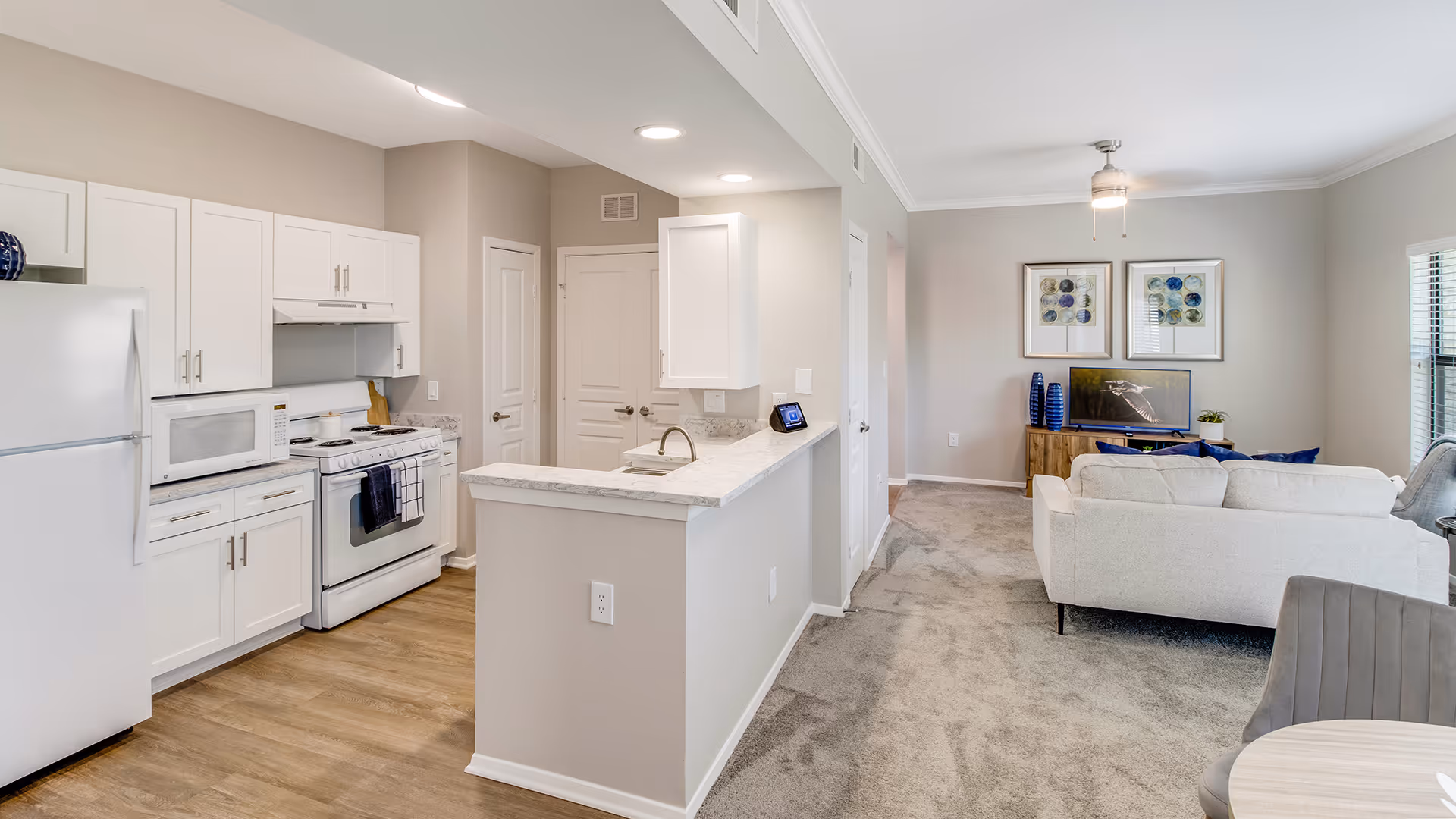 Bright and modern open-concept kitchen and living room area with white cabinets, white appliances including refrigerator, microwave, and stove, a marble countertop with a sink, and a cozy living room with a light-colored sofa, a TV on a wooden stand, decorative blue vases, and framed artwork on the wall.