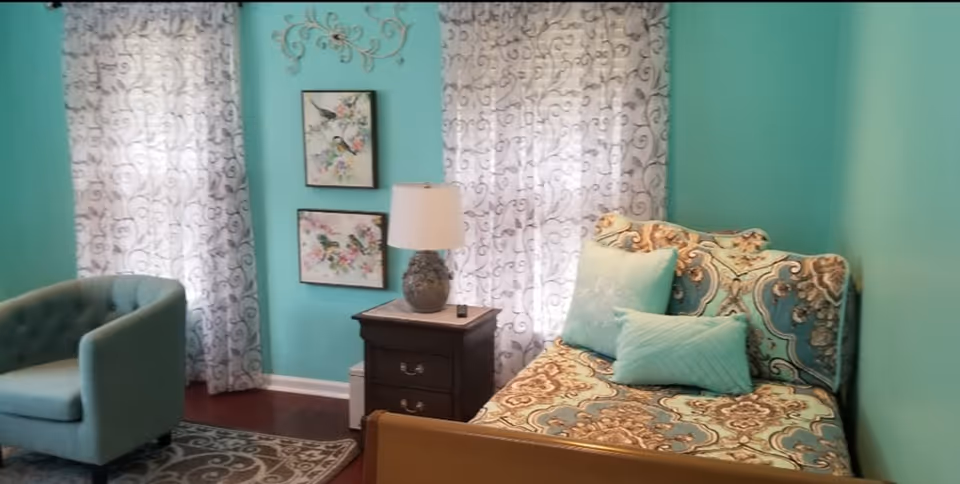 Small turquoise bedroom with a patterned daybed and pillows, a dark nightstand with a lamp, an upholstered chair, and patterned curtains.