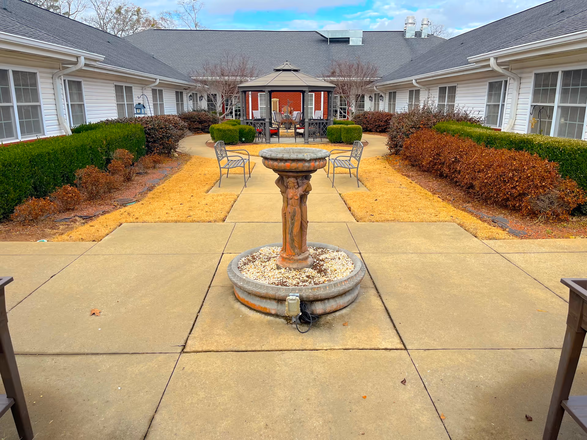 Outdoor courtyard area with a central stone fountain surrounded by a paved walkway. There are metal chairs placed around the fountain and a gazebo with additional seating in the background. The courtyard is bordered by trimmed hedges and bushes, with white buildings on either side under a partly cloudy sky.