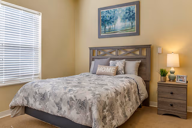 A cozy bedroom with a bed featuring a floral patterned comforter and multiple pillows, including one with the word 'HOME'. There is a wooden nightstand with a lamp, a small plant, and a framed photo. A large window with white blinds allows natural light into the room. A framed painting of trees hangs on the wall above the bed.