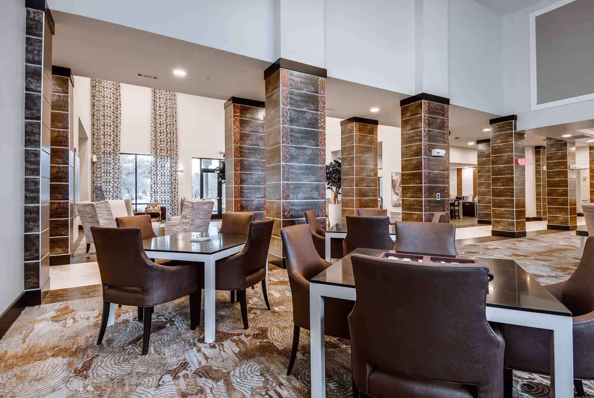 Spacious lounge with multiple square tables and brown leather chairs, tall tiled columns, high ceilings, and patterned carpet.