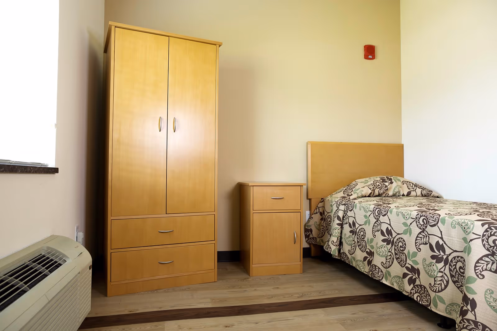 A small single-occupancy bedroom with a light-wood wardrobe, nightstand and a single bed with a patterned bedspread.