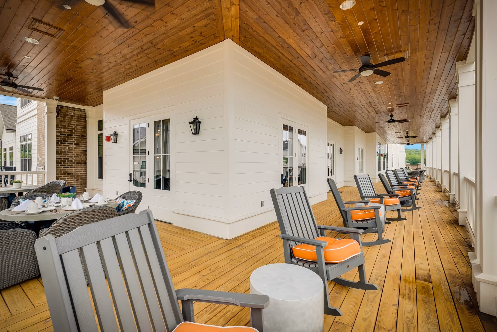 A spacious covered outdoor porch area with wooden flooring and ceiling, featuring a row of gray rocking chairs with orange cushions and small round tables. On the left side, there are round dining tables set with white napkins and wicker chairs. The porch is attached to a white building with multiple doors and windows, and ceiling fans are mounted overhead.