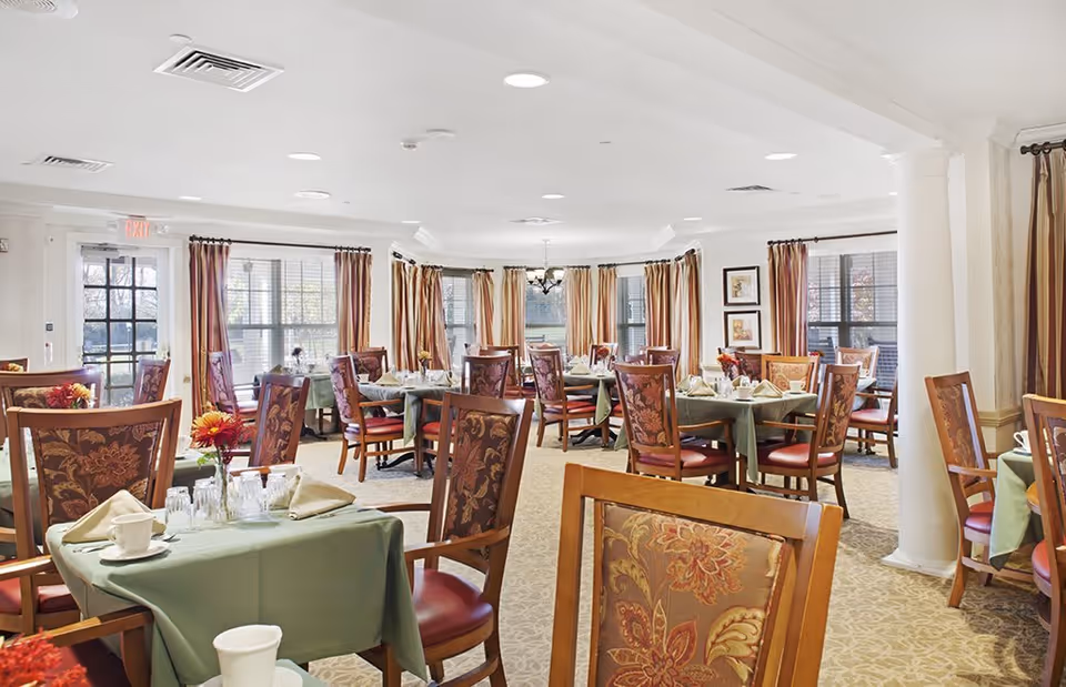 A bright and spacious dining room with multiple tables covered in green tablecloths, each set with napkins, cups, and glasses. The room features large windows with brown and beige curtains, floral upholstered chairs, and a chandelier hanging from the ceiling. The carpet is beige with a subtle pattern, and there are framed pictures on the walls.