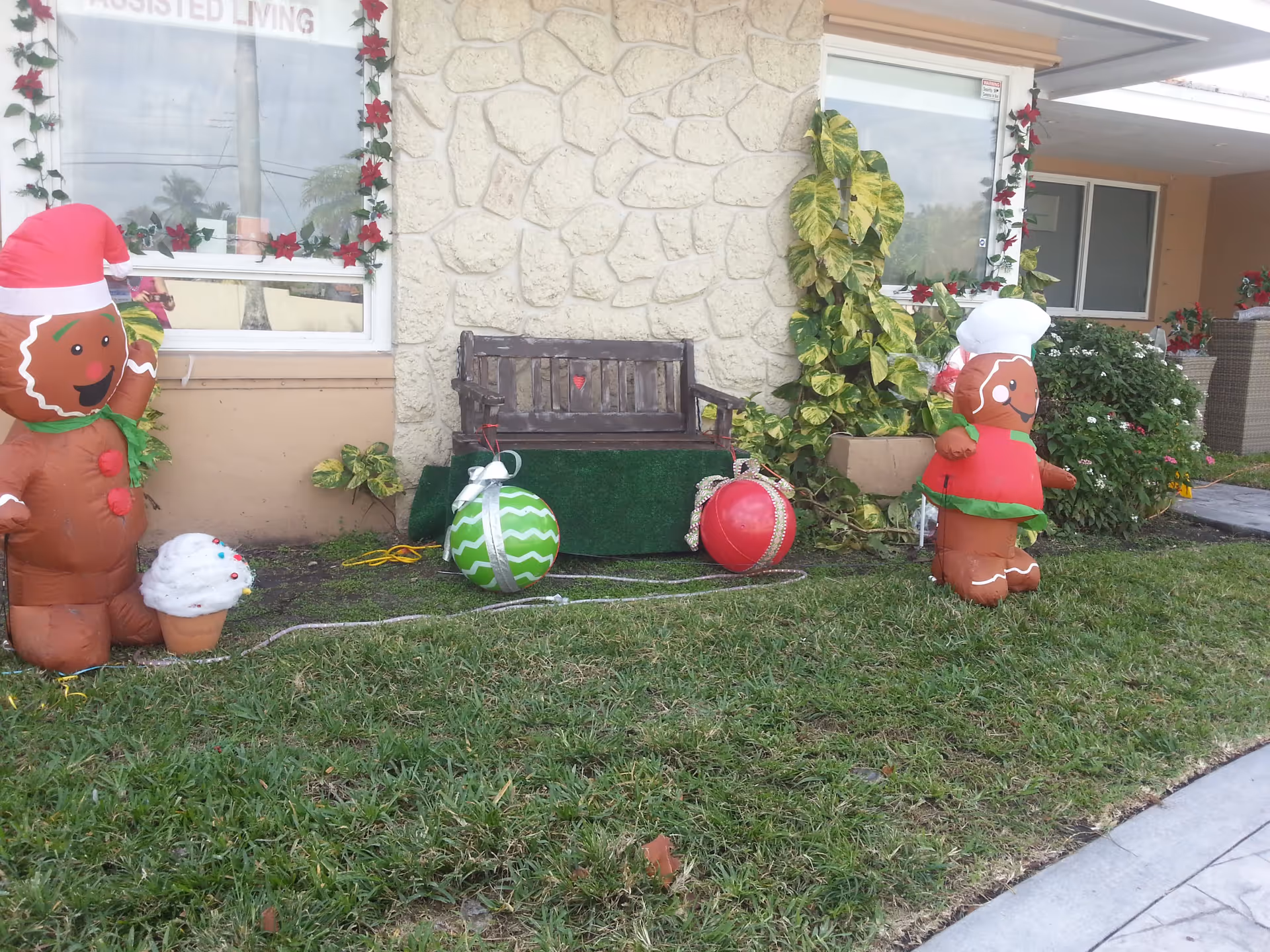 Outdoor front area of Kendall Gardens Assisted Living Facility decorated for the holidays with inflatable gingerbread figures, large Christmas ornaments, a wooden bench, and plants. The building exterior features stone and beige walls with windows framed by garlands of red poinsettias.