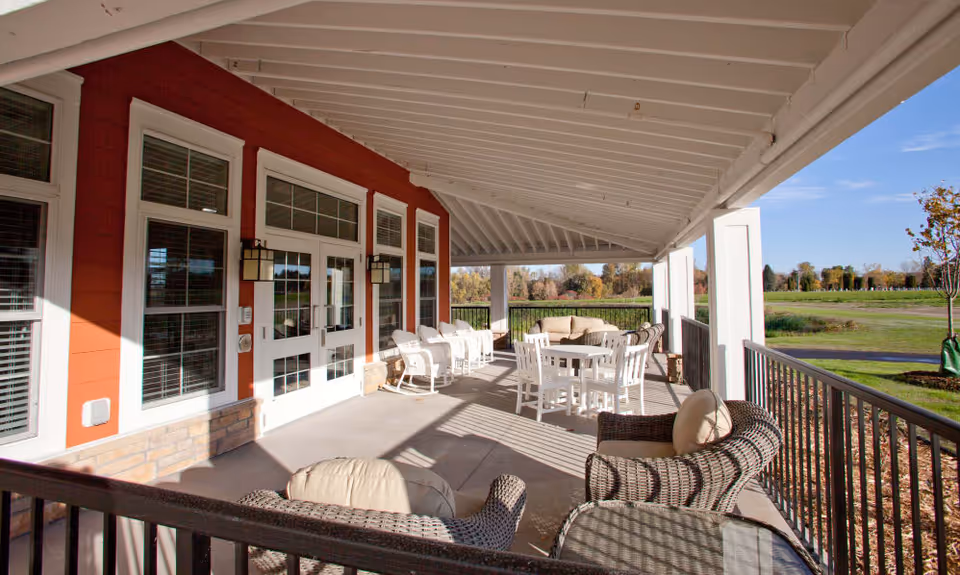 Covered outdoor patio area with wicker chairs, cushioned seating, white rocking chairs, and a white table with chairs. The patio is attached to a red building with large windows and glass doors. Beyond the patio, there is a grassy open field and trees under a clear blue sky.